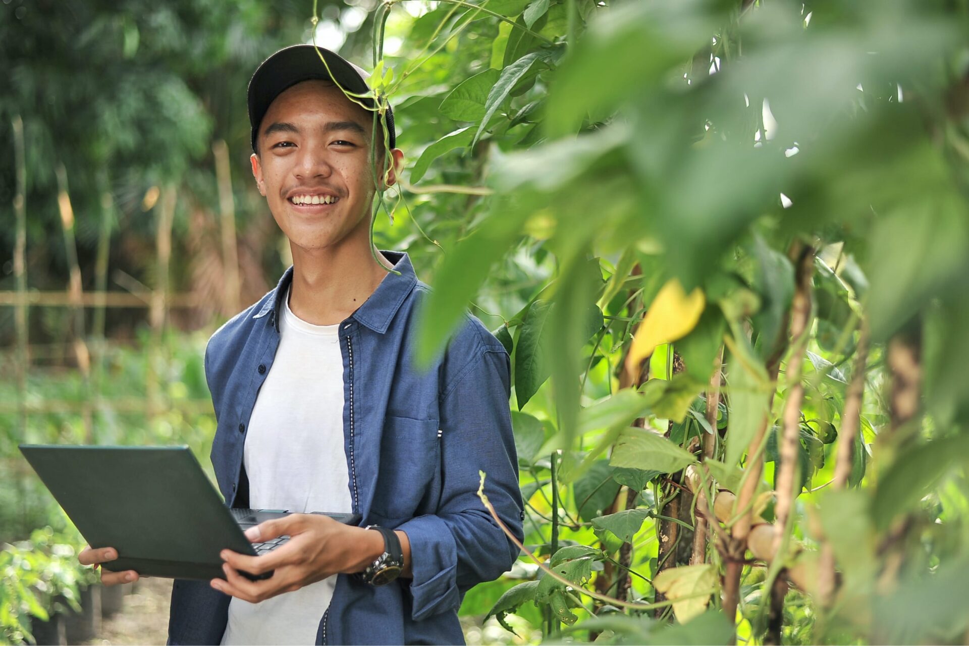 A man standing in field or agriculture setting holding a laptop and smiling into the camera