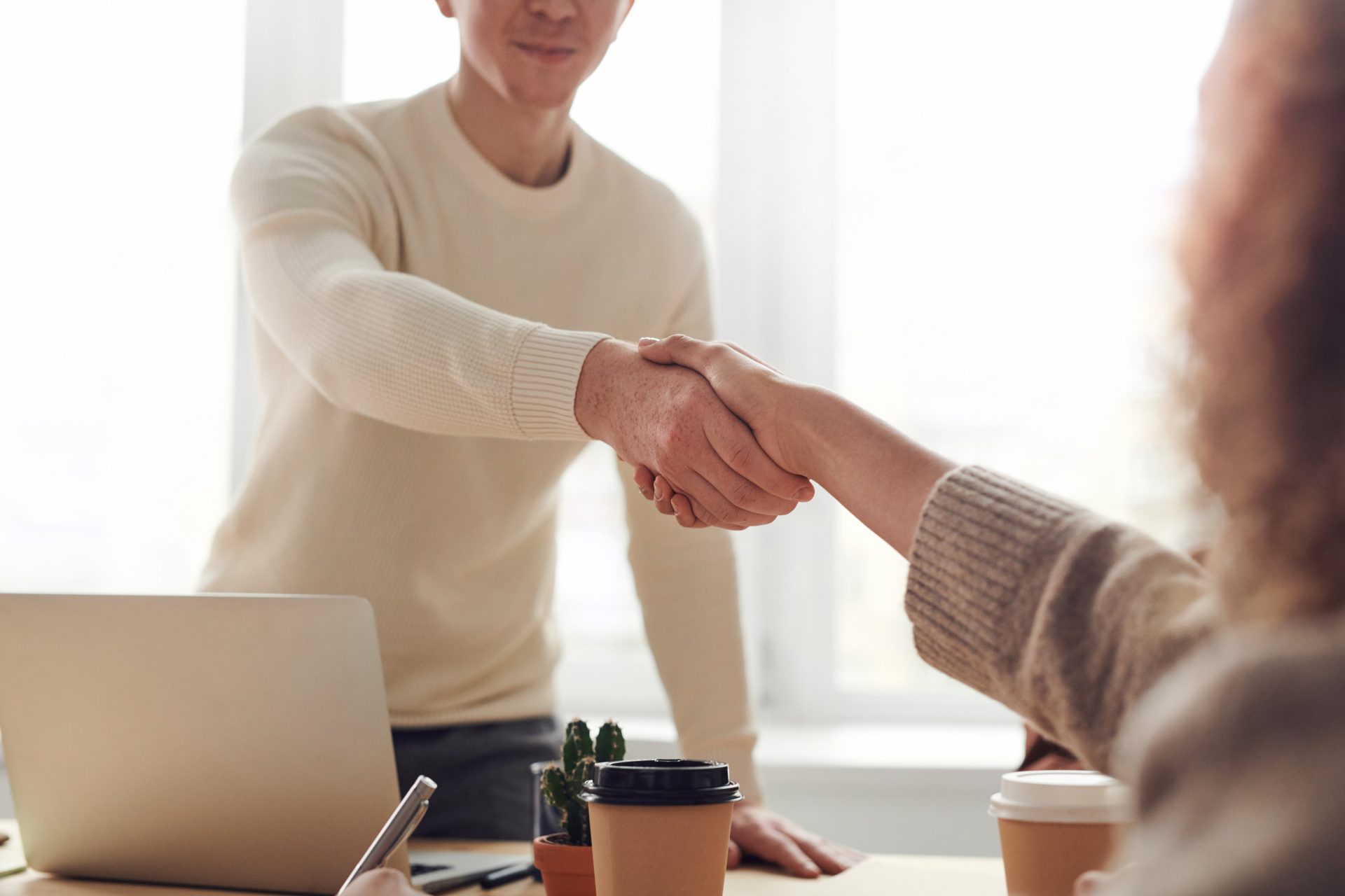 A person standing up in front of a laptop on a desk reaching to shake the hand of another individual